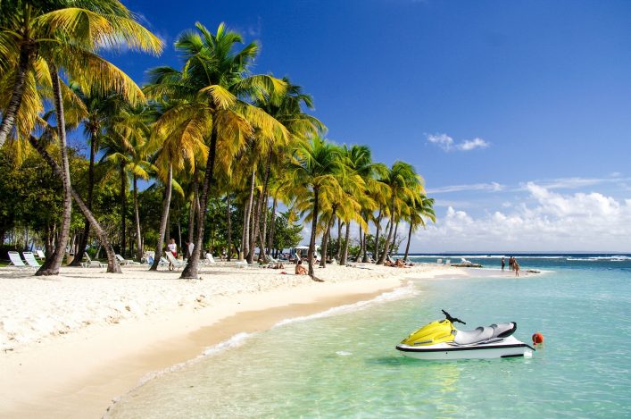 Palmen säumen den Strand, der gegen das türkisfarbene Meer abfällt. Ein gelbes Jetski schwimmt im Vordergrund im Wasser, während Menschen am Strand entspannen und ein Paar spaziert.