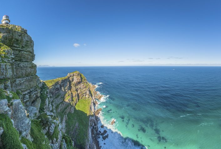Felsige Küste mit grün bewachsenem Hang und Leuchtturm links, darunter türkisblaues Meer, das an die Felsen brandet, im Hintergrund weites, tiefblaues Meer unter klarem Himmel.