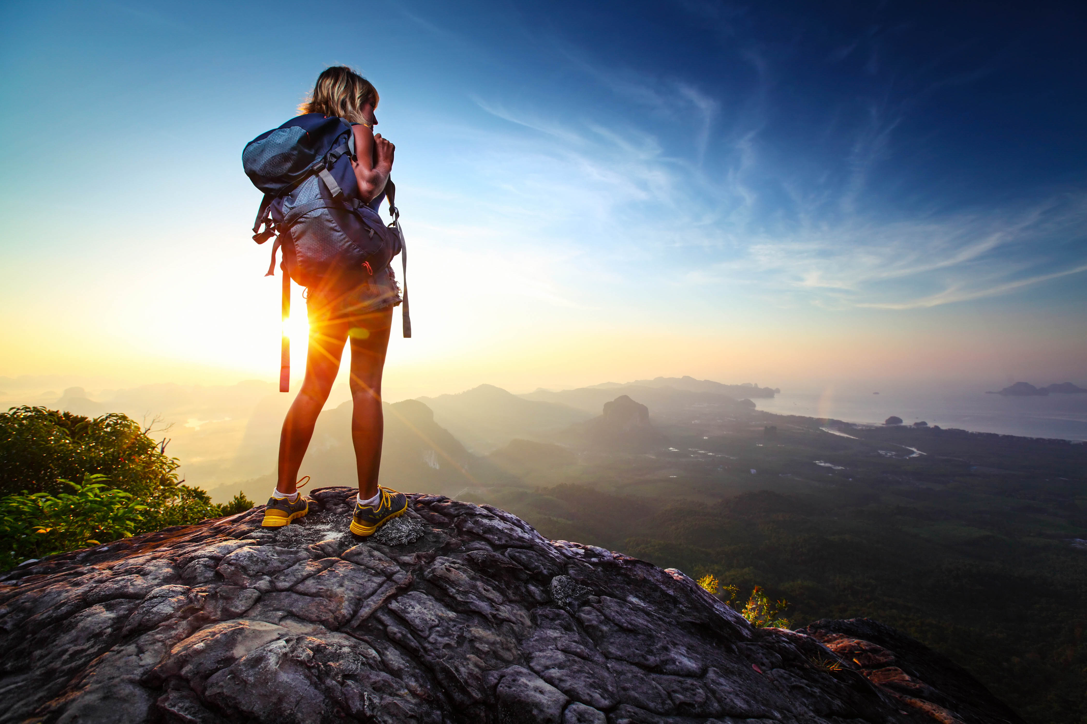 Young lady hiker standing with backpack on top of a mountain and enjoying sunrise