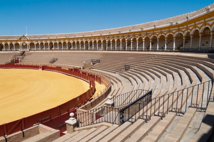 Rundes Stierkampfstadion mit gelber Sandarena in der Mitte, umgeben von mehrreihigen Steinstufen. Im Hintergrund elegante, gebogene Arkaden und klarer blauer Himmel darüber.
