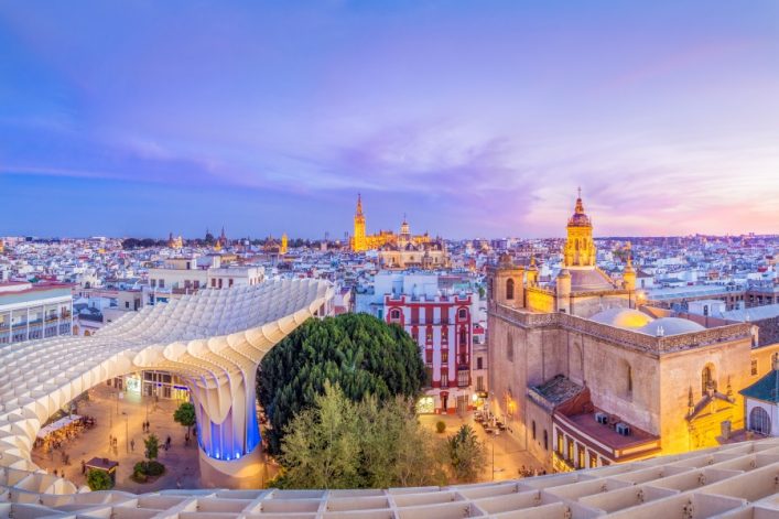 Im Vordergrund erstreckt sich das moderne Holzdach des Metropol Parasol in Sevilla. Rechts steht eine historische Kirche. Im Hintergrund sind beleuchtete Gebäude und ein farbiger Abendhimmel zu sehen.