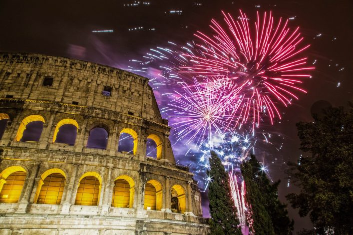 Buntes Feuerwerk erleuchtet den Nachthimmel über dem Kolosseum in Rom, links im Bild. Vorne ragen dunkelgrüne Bäume empor, während der Himmel in leuchtenden Farben strahlt.
