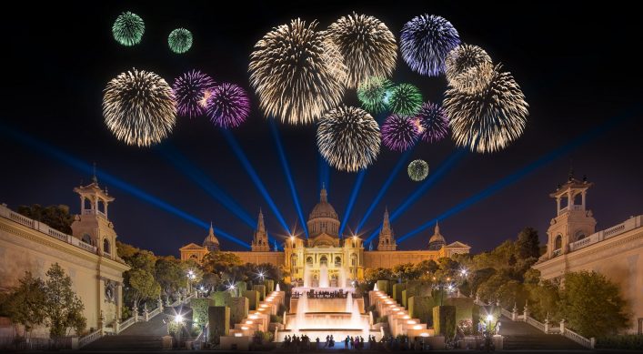 Silvesterfeuerwerk über dem beleuchteten Nationalpalast in Barcelona, umgeben von bunten Feuerwerkskörpern am Himmel, blauen Lichtstrahlen und Menschen vor illuminierten Springbrunnen.