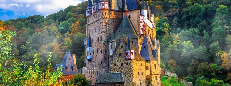 Märchenschloss mit spitzen Türmen auf grünem Hügel, umgeben von dichtem Wald. Blauer Himmel mit weißen Wolken im Hintergrund. Zugang über eine schmale Steinbrücke unten im Bild.