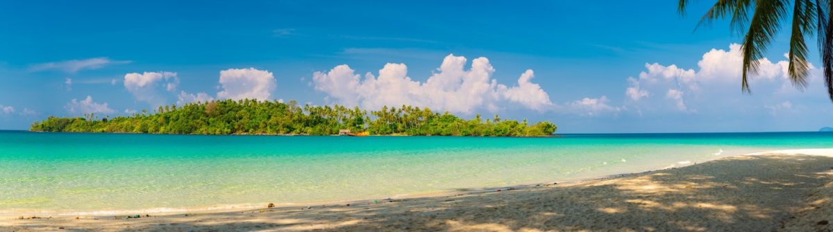 Strand mit hellem Sand im Vordergrund, grüne Palmen rechts. Türkisfarbenes Meer erstreckt sich bis zu einer üppig grünen Insel im Hintergrund. Blauer Himmel mit weißen Wolken darüber.