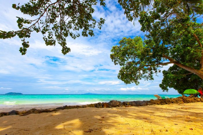 Sandstrand mit Palmen, die Schatten werfen. Rechts zwei Liegen und bunte Sonnenschirme. Türkisfarbenes Meer mit kleinen Wellen, blauer Himmel mit weißen Wolken am Horizont. Im Hintergrund Inseln.