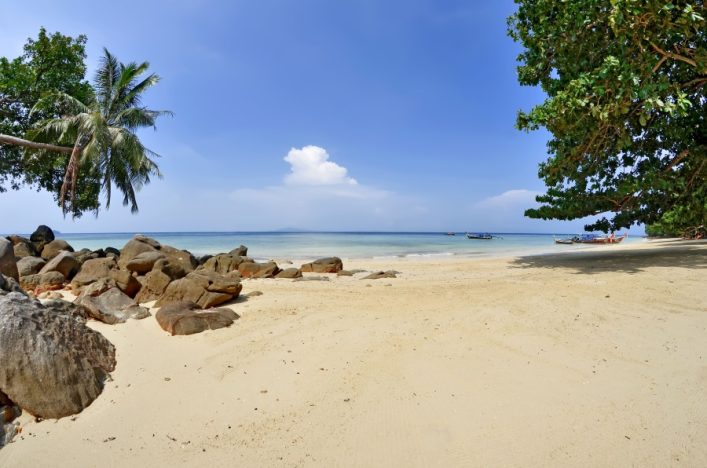 Ein sonniger Strand mit hellem Sand, links Felsen und Palmen, rechts dichte grüne Bäume. Im Hintergrund das klare blaue Meer mit mehreren kleinen Booten unter einem leicht bewölkten Himmel.