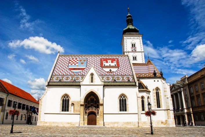 Die Markuskirche in Zagreb von vorne fotografiert. Sie ist weiß und hat auf den Dächern kroatische Wappen und andere Muster aus den Farben des Landes.