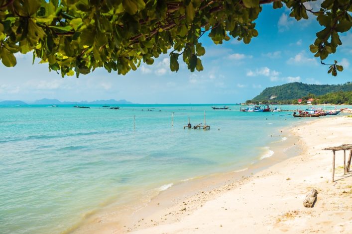 Am Thong Krut Strand leuchtet türkisblaues Wasser unter klarem Himmel. Boote schwimmen nahe der Küste, die von grünen Bäumen umrahmt ist. Der Sand verläuft sanft ins Wasser.
