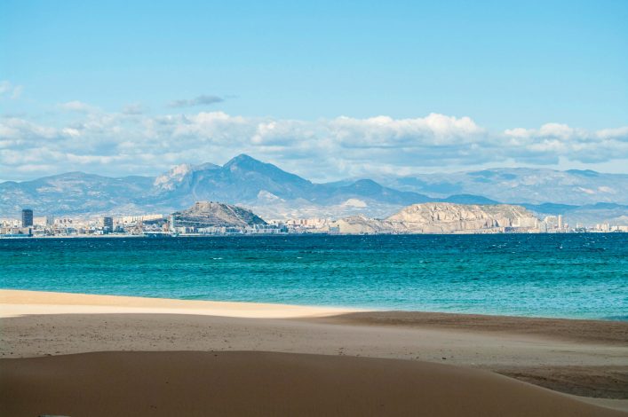 Klarer Himmel über Alicante; im Vordergrund ein Sandstrand, dahinter türkisfarbenes Meer; die Küstenlinie und die Stadt im Hintergrund sind vor einer Bergkette sichtbar.