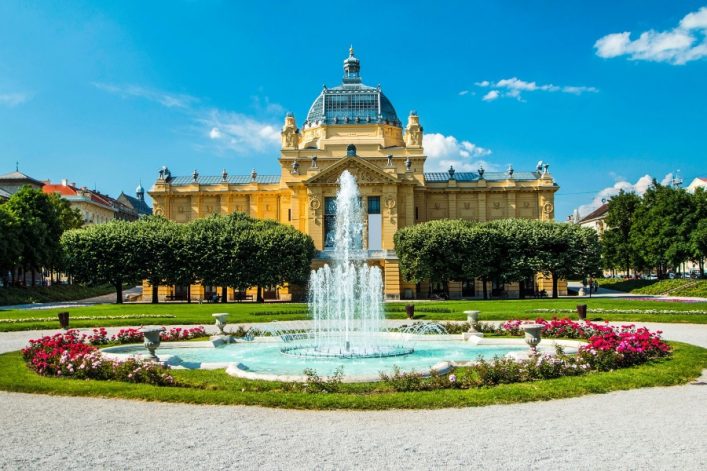 Man sieht das Nationaltheater von Zagreb. Davor einen Brunnen, der gerade Wasser wirft. Um ihn herum sind Blumenbeete. Der Himmel und das Wasser sind hellblau.