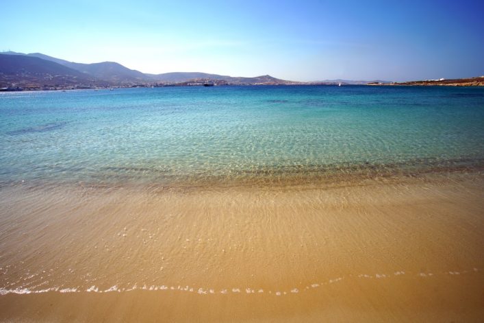Feiner Sandstrand führt zu türkisfarbenem, klarem Wasser, das sich sanft gegen den Ufer spült. Im Hintergrund erstrecken sich sanfte Hügel und der Himmel ist klar und blau.
