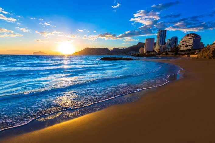 Sonnenuntergang am Strand von Calpe, Spanien: Links die goldene Sonne über dem Meer, rechts moderne Hochhäuser am Ufer. Wellen treffen auf den Sand, sanfte Wolken färben sich am Himmel bunt.