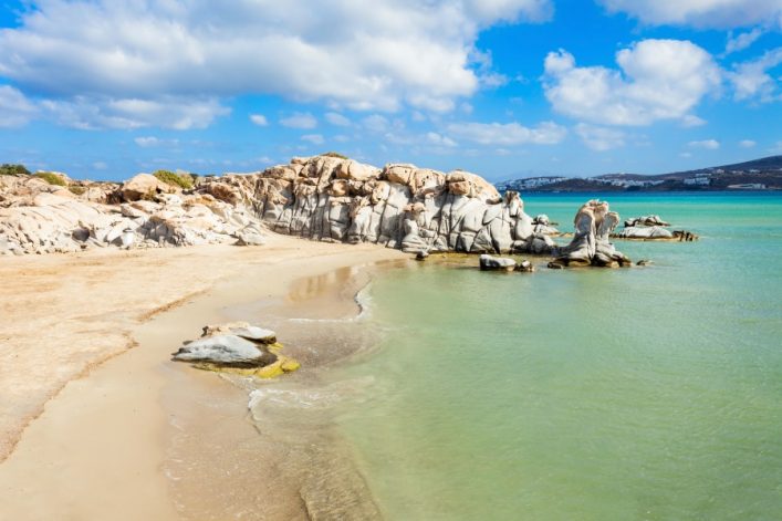 Sandstrand und türkisfarbenes Wasser umgeben von bizarren Felsformationen am Kolimbithres Beach. Felsen dominieren rechts, während links der Sand sanft ins Meer übergeht. Im Hintergrund blauer Himmel mit Wolken.
