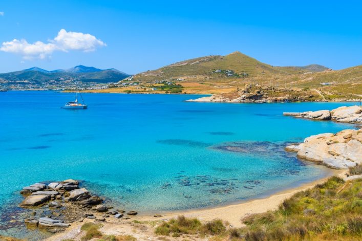 Klares türkisfarbenes Meer mit einem Boot in der Mitte, Sandstrand und Felsen im Vordergrund, grüne Hügel und weiße Gebäude im Hintergrund unter blauem Himmel mit ein paar Wolken.