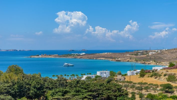 Türkisblaues Meer umrahmt eine felsige Küste mit vereinzelten weißen Häusern. Im Vordergrund grüne Bäume, im Hintergrund sanfte Hügel und wolkenfreier blauer Himmel. Eine Yacht schwimmt im Wasser.