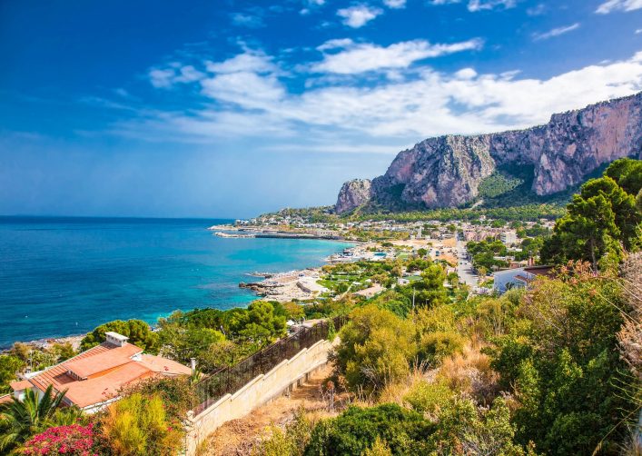 Panoramablick auf die Bucht von Mondello in Palermo, Sizilien: Klares Meer im Vordergrund, Küstenstadt und üppige Berge dahinter unter blauem Himmel.