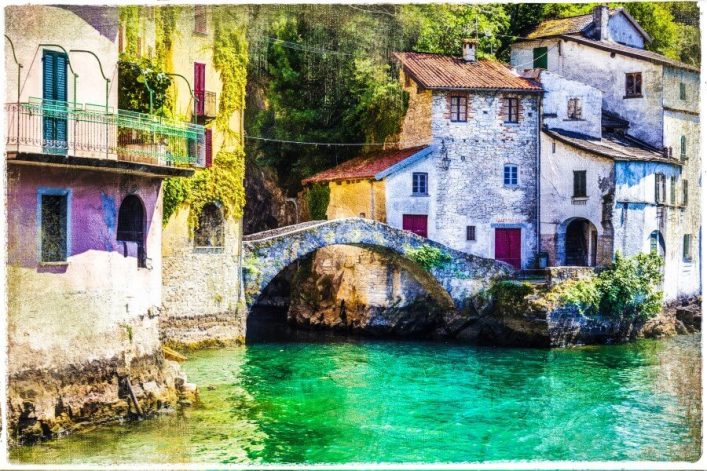 Malerische Häuser mit roten Dächern stehen an einem smaragdgrünen See. Eine steinerne Brücke verbindet die Gebäude, die von Efeu umgeben sind. Helle Farben betonen die Idylle der Landschaft.