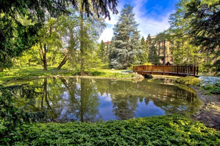 Der Botanische Garten von Zagreb. Zu sehen sind ein grünlicher See und drumherum viele Bäume und Sträucher. Rechts auf dem Bild ist eine braune kleine Brücke zu sehen.