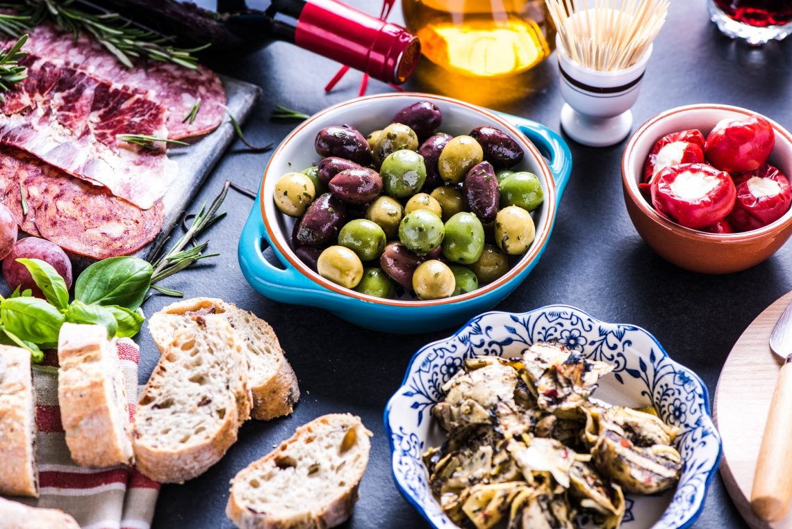 Verschiedene spanische Tapas auf einem Tisch: grüne und schwarze Oliven in der Mitte, gefüllte Paprika rechts, links Brot, darüber Salamischeiben und Rosmarinzweige, im Vordergrund Artischocken.