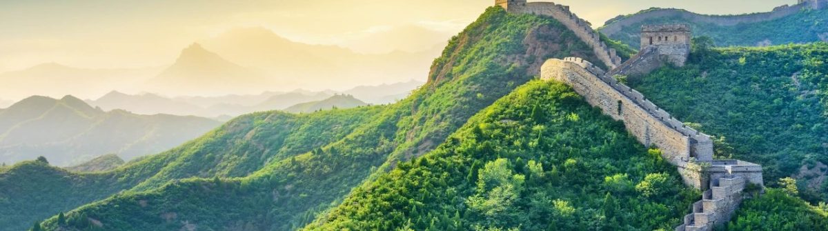 Teil der Chinesischen Mauer schlängelt sich über grüne Berge unter einem blauen Himmel mit aufgehender Sonne, ein Weltwunder der Antike.