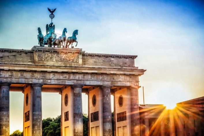 Brandenburger Tor mit der Quadriga oben, von Säulen getragen. Goldener Sonnenuntergang rechts erzeugt helle Strahlen im Bild, mit blauem Himmel im Hintergrund.