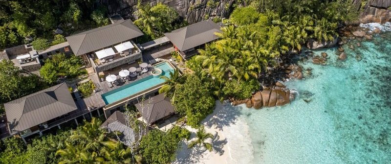 Luxuriöse Strandvillen mit Pool liegen zwischen üppigem Grün und Felsen, daneben ein weißer Sandstrand mit klarem, türkisfarbenem Wasser. Palmen säumen den Rand der Villa, die naturverbunden wirkt.