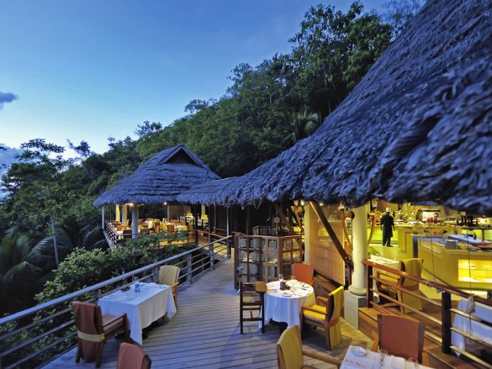 Holzterrasse eines Restaurants mit Strohdach, eingerahmt von grüner Vegetation; links Sitzgelegenheiten und gedeckte Tische, rechts Buffetbereich mit gelbem Licht; blauer Himmel in der Dämmerung.