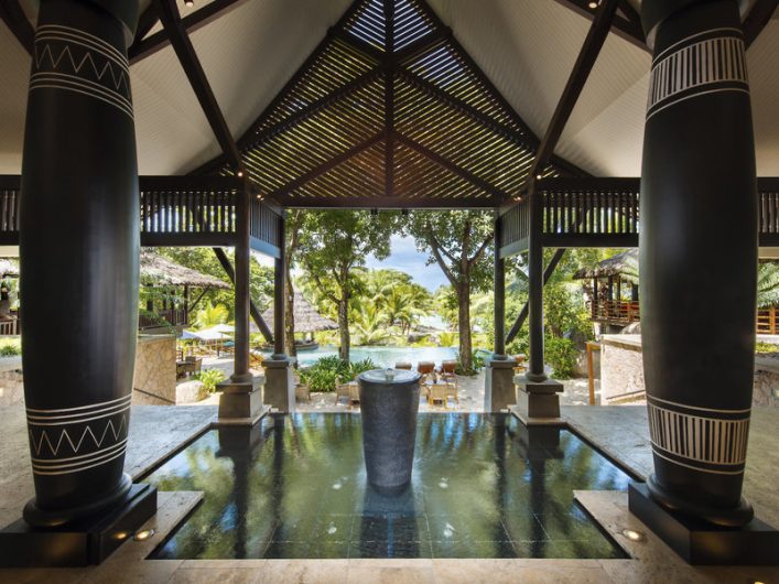 Hölzerner Pavillon mit zwei großen, verzierten Säulen seitlich. Ein Wasserbecken mit Brunnen steht zentral im Vordergrund. Palmen und tropische Vegetation sind im Hintergrund sichtbar.