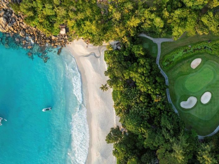 Strand mit weißem Sand liegt zwischen türkisfarbenem Meer links und dichter grüner Vegetation rechts. Ein Golfplatz mit zwei Sandbunkern ist oben rechts sichtbar. Ein Boot schwimmt im Meer.