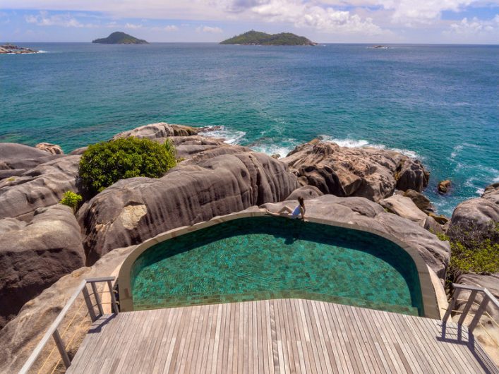 Ein infinity-pool über blickt das türkisfarbene Meer, umgeben von mächtigen grauen Felsen. Im Hintergrund sind zwei kleine Inseln. Eine Person steht im Pool, eine Holzterrasse liegt im Vordergrund.