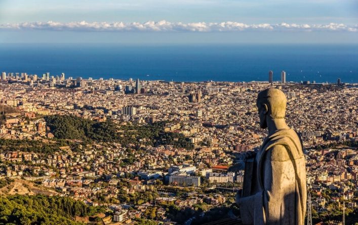 Panoramablick auf Barcelona mit einer Statue im Vordergrund, dichtbebauter Stadtfläche und Meer im Hintergrund bei Tageslicht.
