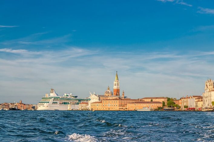 Ein großes Kreuzfahrtschiff ankert nahe der Küstenlinie Venedigs, hinter dem die Silhouette der Stadt mit markanten Türmen zu erkennen ist, unter blauem Himmel.