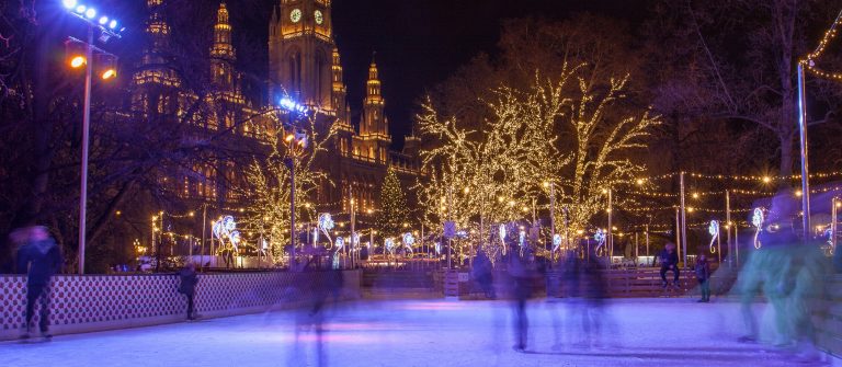 Beschneite Bäume schimmern im Lichterglanz neben einer Eislaufbahn in Wien, vor dem illuminierten Rathaus. Menschen genießen das nächtliche Schlittschuhlaufen.