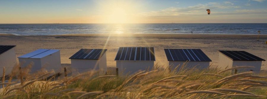 Sonnenuntergang am Strand in der Provinz Zeeland, Niederlande. Im Vordergrund Dünengras, dahinter weiße Strandhütten und dazwischen ein Kite-Surfer in der Ferne über dem Meer.
