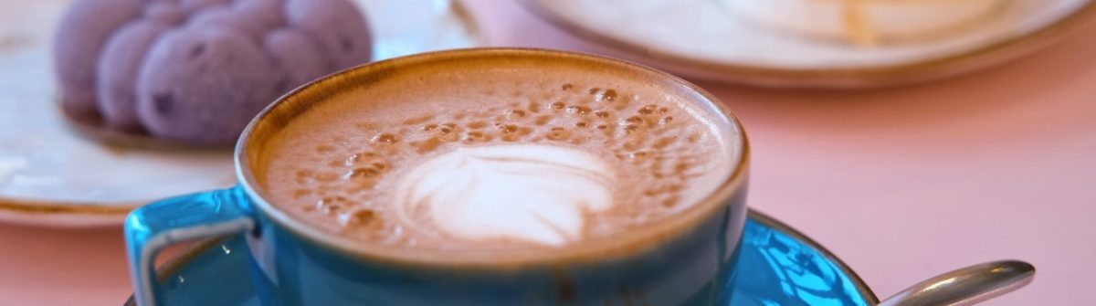 In einem der besten Cafés: Links im Vordergrund ein Cappuccino in einer blauen Tasse mit Schaum, rechts ein Teller mit einem Stück Torte und im Hintergrund violette Macarons.