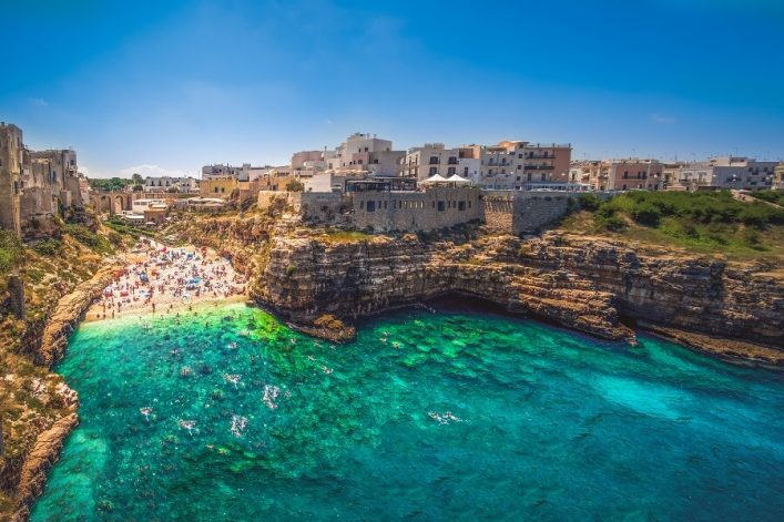 Klippenbucht in Apulien mit klarem türkisblauem Wasser, Menschen sonnen sich auf Felsen, umgrenzt von historischen Gebäuden.