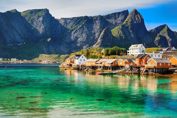 Ein Bild der Lofoten. In der unteren Hälfte ist türkis-grünes Wasser. In der oberen Hälfte ragen kahle Berge in den Himmel. Auf der rechten Seite des Bildes liegen bunte Häuser am Wasser.