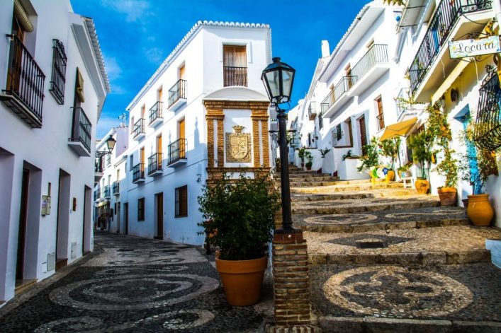 Pflasterstraße in Frigiliana, Spanien, gesäumt von weiß gestrichenen Häusern mit Balkonen. Rechts führen Treppen nach oben, Pflanzen in Töpfen stehen verteilt, eine Laterne in der Mitte.