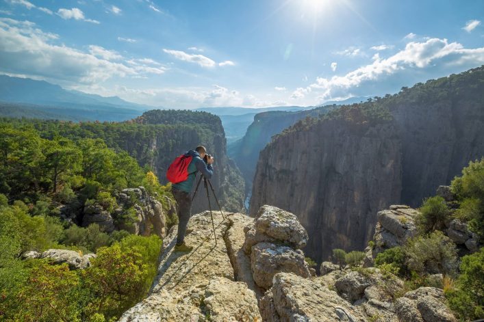 Ein Fotograf mit rotem Rucksack steht links auf einem Felsen und fotografiert eine spektakuläre Berglandschaft. Rechts und im Hintergrund erstrecken sich bewaldete Schluchten unter blauem Himmel.