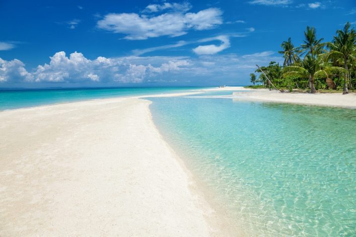 Türkises Wasser umspült eine weiße Sandbank, die in den Horizont verläuft. Rechts Palmen am Strand, der Himmel strahlend blau mit wenigen Wolken, vermittelt ein Gefühl von tropischer Idylle.