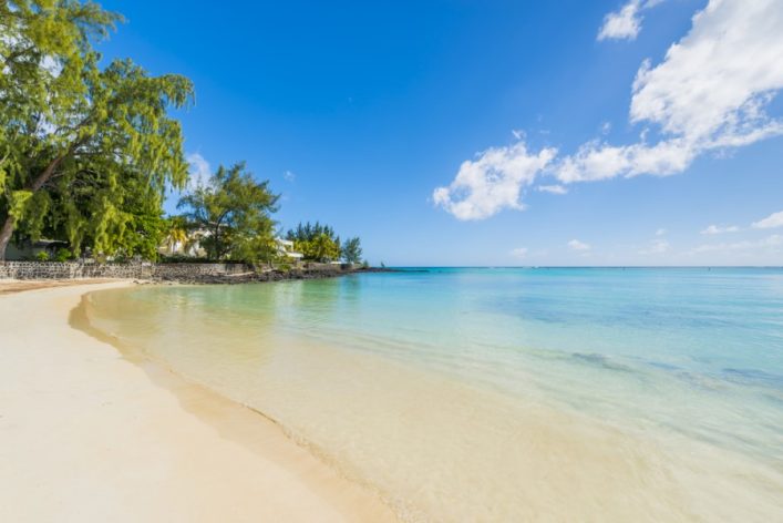 Feiner Sandstrand im Vordergrund, der sanft ins türkisfarbene Meer übergeht. Links säumen grüne Bäume die Küstenlinie, die sich bis zum Horizont erstreckt. Blauer Himmel mit einigen Wolken.