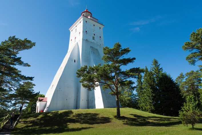 Ein weißer Leuchtturm mit rotem Dach ragt in die Höhe, umgeben von grünen Bäumen und strahlendem blauen Himmel. Im Vordergrund erstreckt sich eine grüne Wiese.