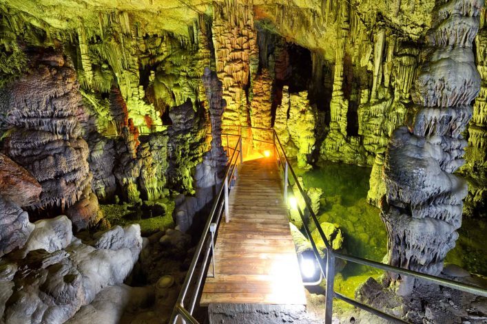 Eine beleuchtete Holzbrücke führt durch die Tropfsteinhöhle von Psychro auf Kreta, umgeben von grün schimmernden Wasserpools und faszinierenden Stalaktiten und Stalagmiten.