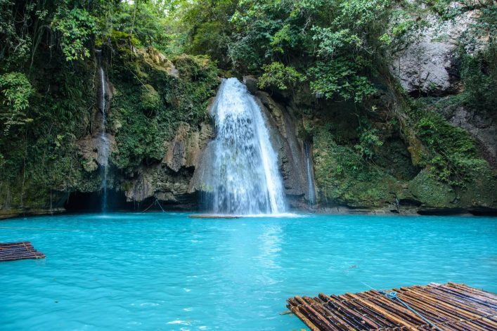 Türkisfarbener Pool mit Wasserfall, der von einer bewachsenen Felswand in der Bildmitte herabstürzt. Dichte Vegetation oben und an den Seiten. Im Vordergrund treiben Bambusflöße auf dem Wasser.