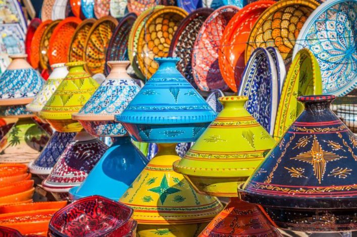 Bunte, handbemalte Keramikwaren stapeln sich auf einem Markt in Marrakesch, darunter Schalen und Tajine-Töpfe in leuchtenden Farben und Mustern.