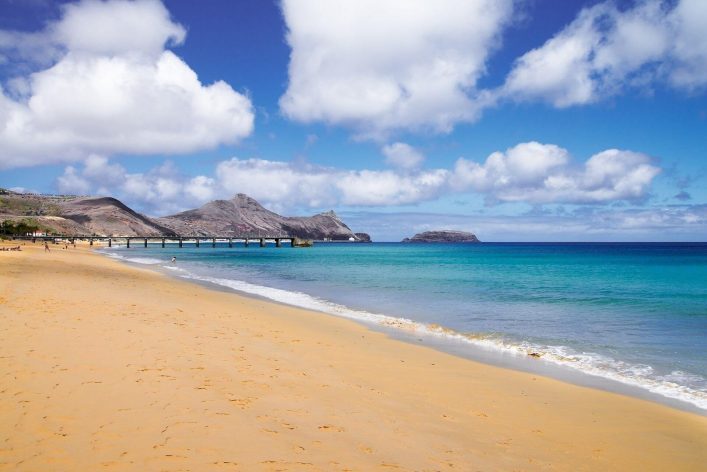 Sandstrand mit sanften Wellen, blauer Himmel und wenigen Wolken. Rechts das azurblaue Meer, links eine Holzbrücke. Im Hintergrund felsige Hügel. Die Szene strahlt Ruhe und Naturschönheit aus.