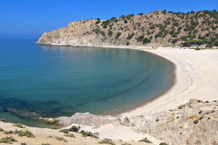 Halbmondförmige Bucht mit Sandstrand; kristallklares, türkisfarbenes Wasser. Links felsige Klippen, rechts bewaldete Hügel. Leichter Wellengang am Ufer, blauer Himmel darüber.