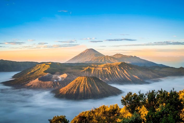 Sonnenaufgang über dem Mount Bromo mit leuchtend rot-orange gefärbtem Krater in der Mitte, umgeben von Nebel. Im Hintergrund erhebt sich ein anderer Vulkan unter klarem, blauem Himmel.