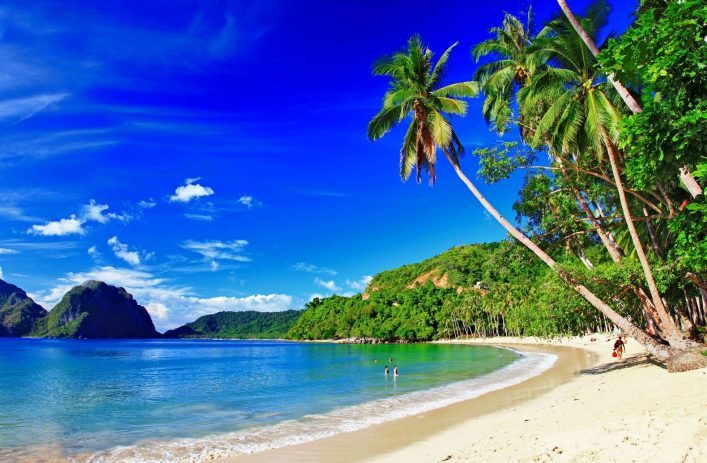 Sandstrand von El Nido mit türkisfarbenem Wasser und blauem Himmel, links Hügel im Meer, rechts Palmen und dichter Wald, Person am Strand und zwei im Wasser.